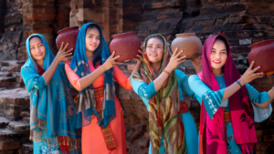 4 smiling women in bright dresses and scarves hold clay plots over their left shoulders