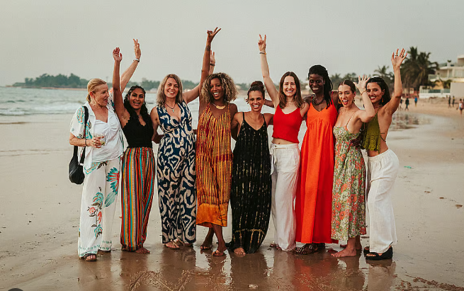 RETREATIMG_2 9 smiling women pose for a photo on a wet sandy beach, each raising 1 arm in the air with their fingers in peace signs.
