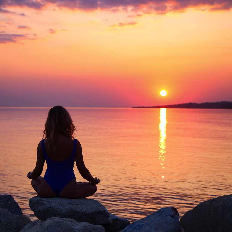 A woman sits cross-legged on a rock overlooking a calm ocean as the sun rises in the distance.