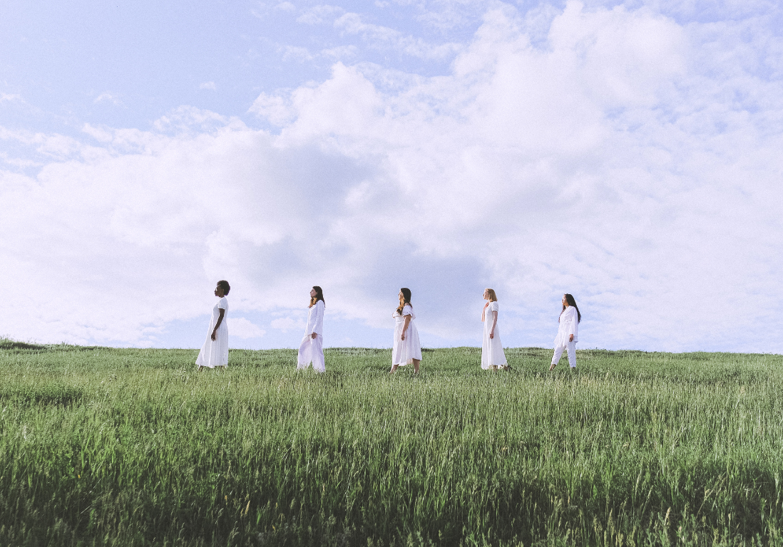 11 5 women wearing white walking in a line through a field of tall green grass.