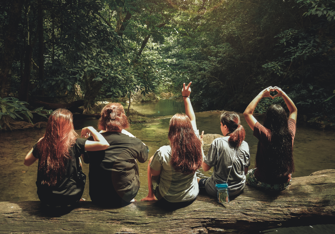 20 5 women sit on a log over a shallow creek with their backs to the camera, looking into the depths of a forest.