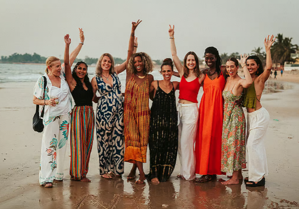 9 smiling women pose for a photo on a wet sandy beach, each raising 1 arm in the air with their fingers in peace signs.
